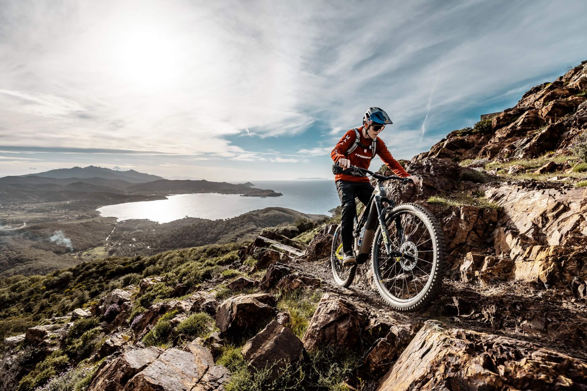 Mountainbiker auf Haibike fährt steinigen Hang hinunter, im Hintergrund eine Landschaft mit See und Bergen bei Sonnenschein.