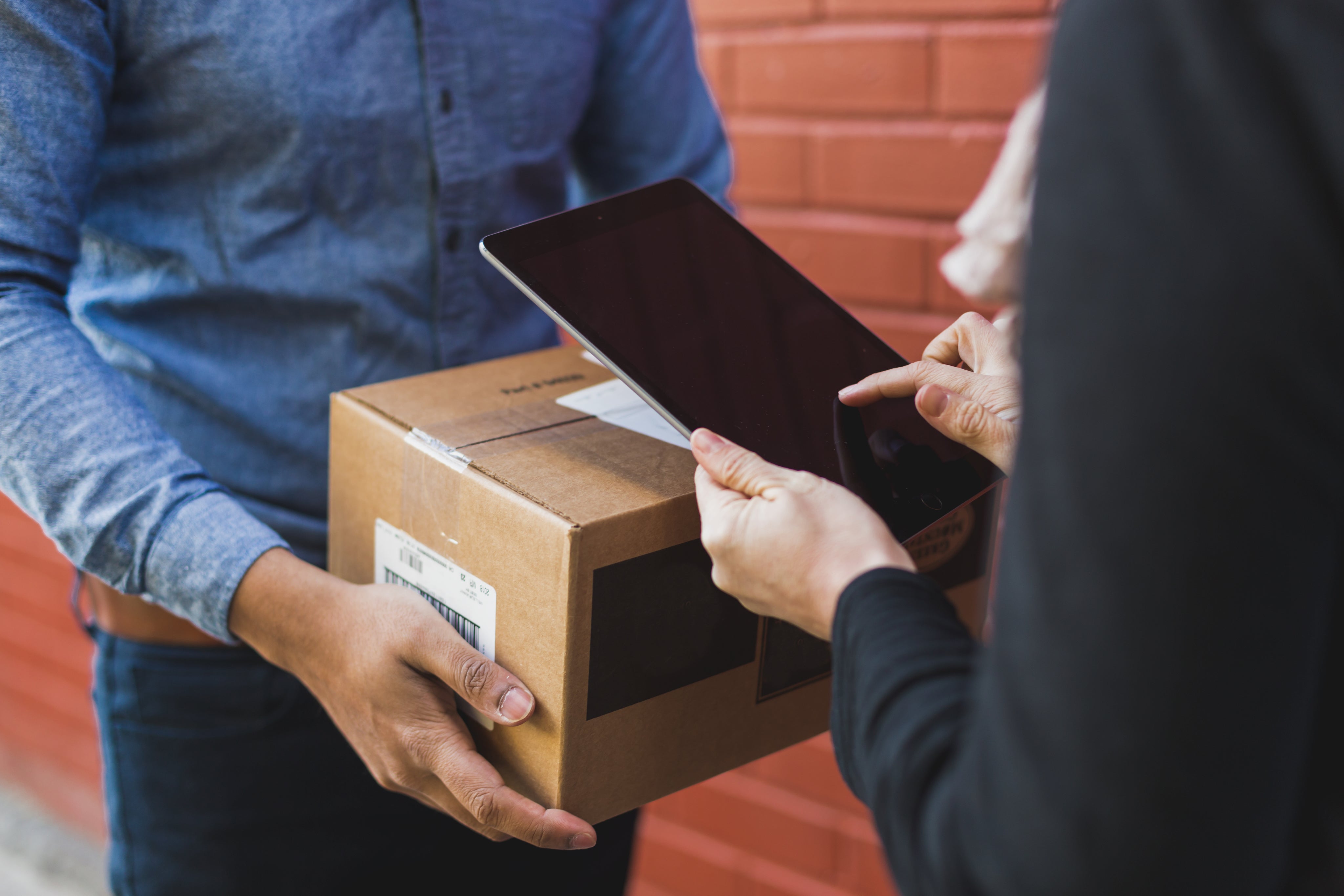 Person erhält Paket, unterschreibt auf einem Tablet, vor einer roten Ziegelwand. Ideal für Lieferdienste.