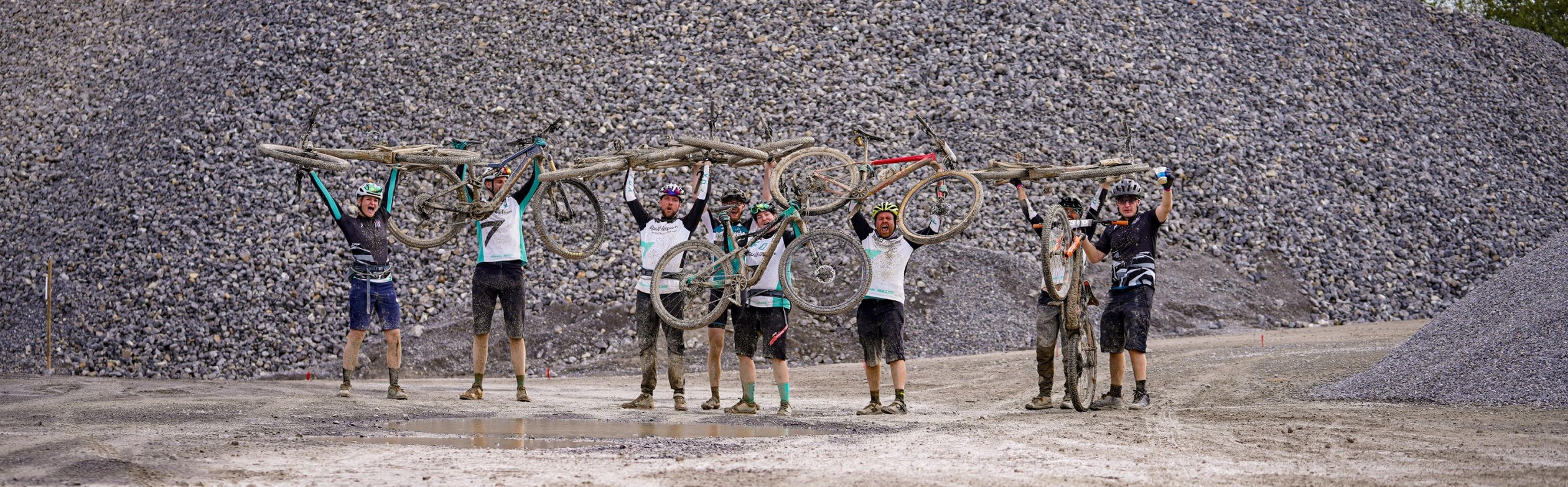 Gruppe von Mountainbikern hält schlammige Fahrräder hoch, vor einem großen Kieshaufen stehend.