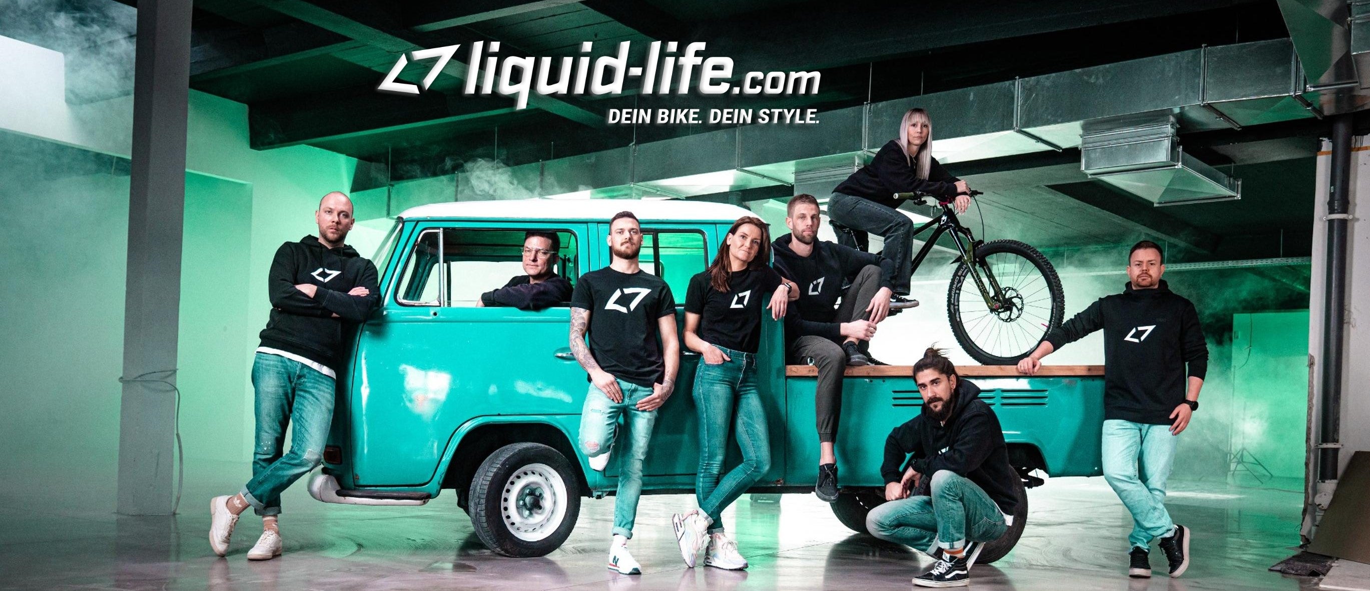 Team von liquid-life.com posiert in Freizeitkleidung mit Van und Fahrrad in industrieller Umgebung.