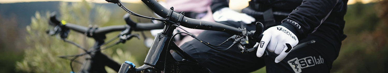Zwei Personen mit Fahrrädern tragen SQlab-Bekleidung und weiße Fahrradhandschuhe, Fahrt durch Naturgebiet.