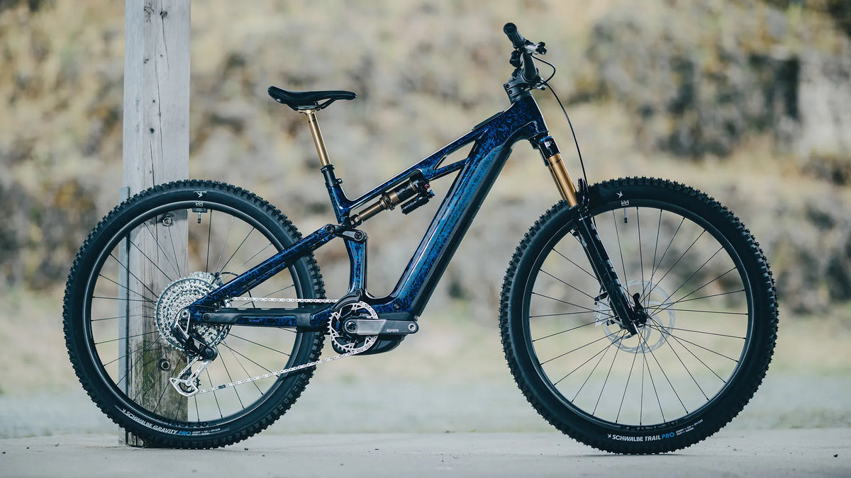 Blaues CUBE STEREO HYBRID ONE44 HPC SLT 800 mit Vollfederung, geeignet für den Geländeeinsatz, abgestellt an einem Holzbalken. Schwalbe-Reifen.