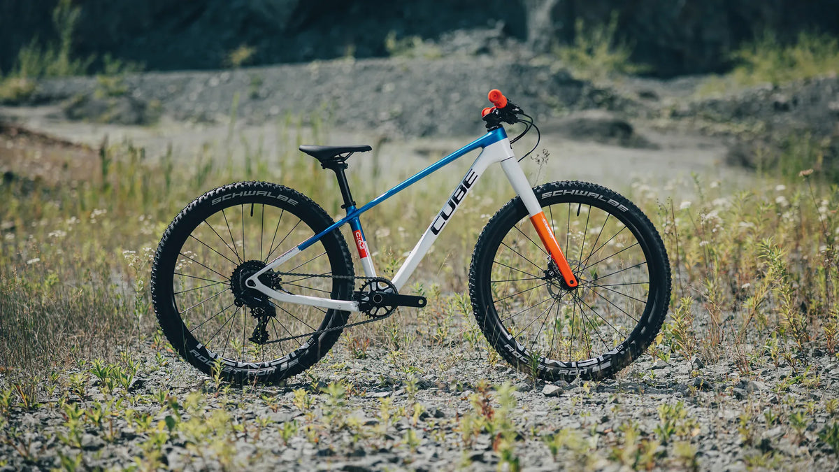Cube PHENIX 240 C:62 SLX 2027 Modell für Kinder, in blau-weiß-orange mit Schwalbe Reifen, auf einem kiesigen Feldweg positioniert.