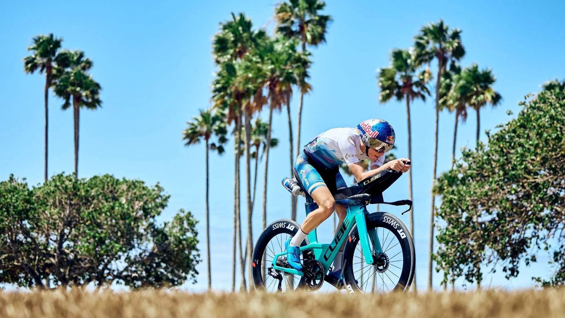 Radfahrer auf blauem Cube Aerium unter Palmen, trägt Sporthelm; ideal für Triathlon, sportliche Zielgruppe.