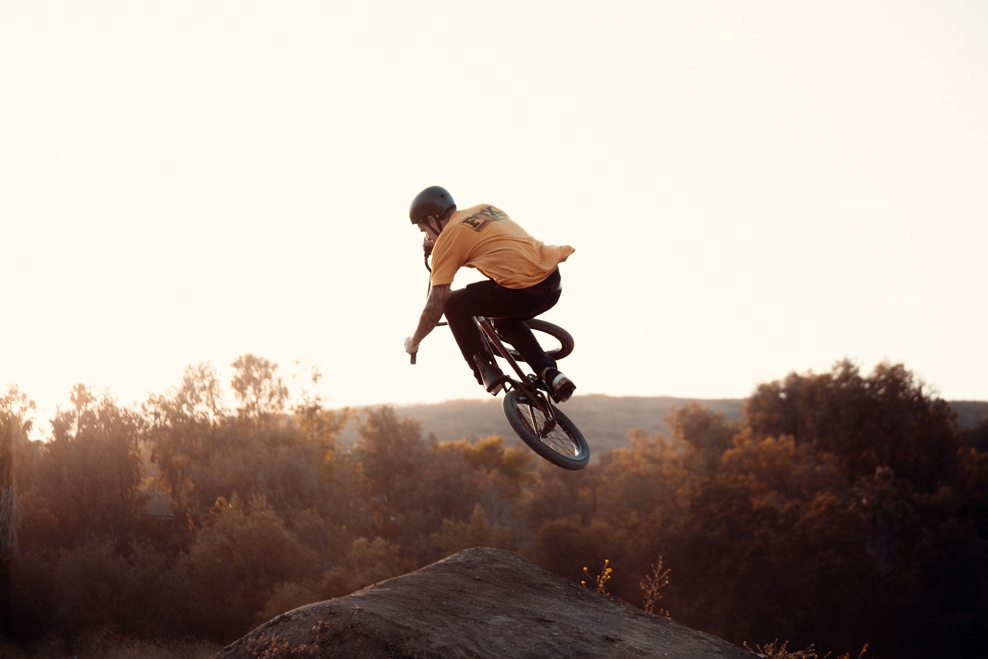 Mensch auf BMX-Rad springt über Hügel, Sonnenuntergang im Hintergrund, trägt Helm und gelbes T-Shirt.