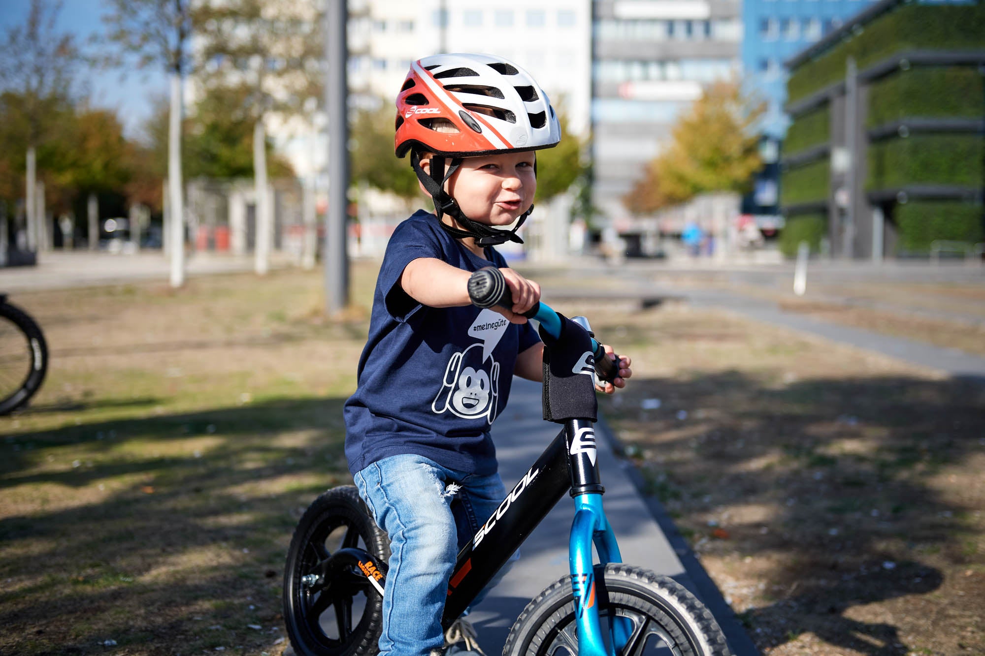 Junge auf einem S'COOL-Laufrad mit rotem Helm, ideal für Kinder von 1 bis 3 Jahren, in städtischer Umgebung.