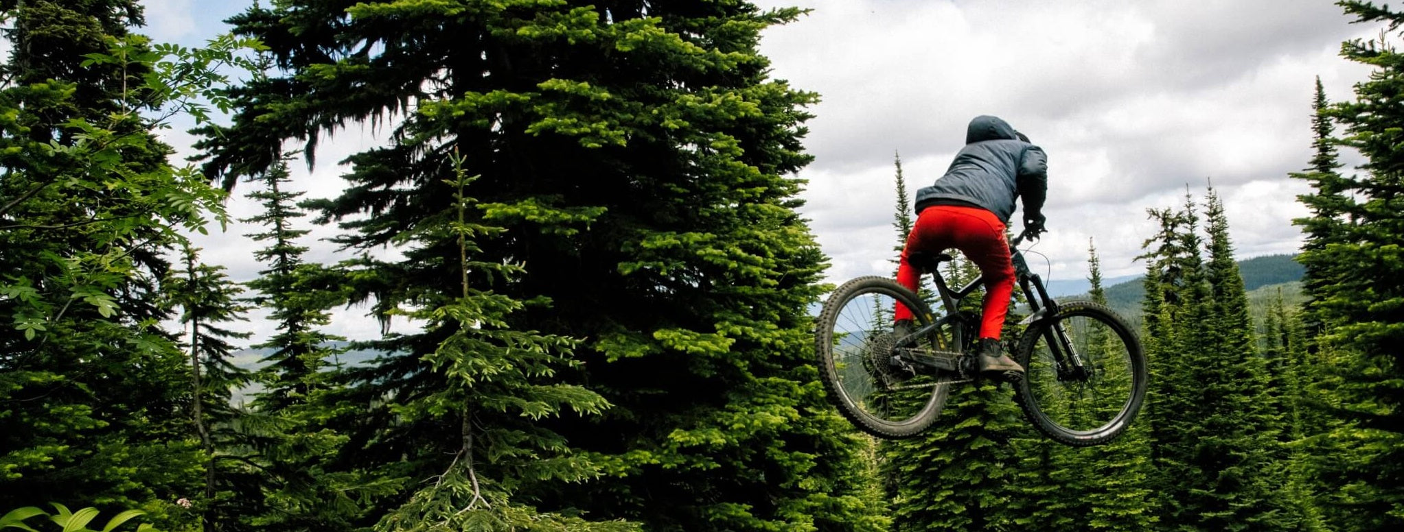 Mountainbiker springt im Wald, trägt rote Hose und dunkle Jacke. Ideal für Offroad-Abenteuer und Outdoor-Enthusiasten.