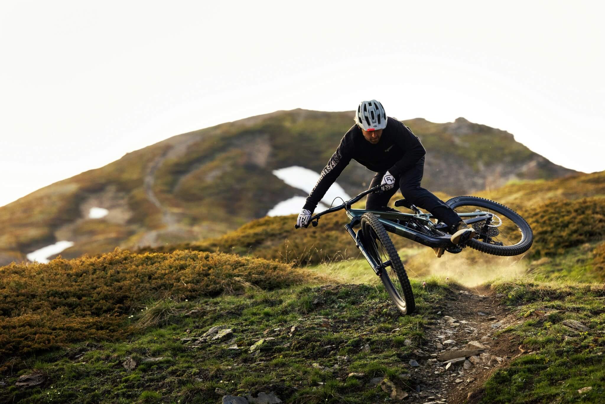 Mountainbike-Fahrer mit schwarzem Fahrrad auf steinigem Bergpfad, dynamische Pose in alpiner Landschaft.