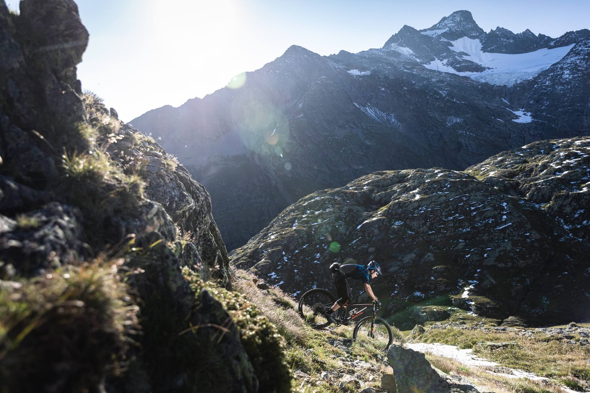 Mountainbiker auf BMC Speedfox fährt steilen, felsigen Weg in den Alpen hinab, Sonne strahlt über die Berggipfel.