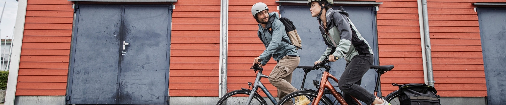 Zwei Personen in Outdoor-Kleidung fahren Fahrräder vor einer roten Holzwand. Beide tragen Helme und Rucksäcke.