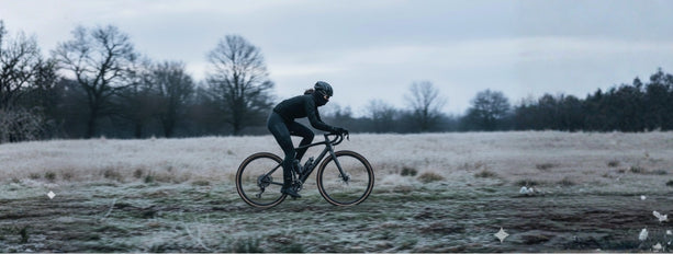 Radfahrer auf schwarzem Fahrrad in Winterkleidung, fährt durch eine frostige Landschaft mit kahlen Bäumen im Hintergrund.