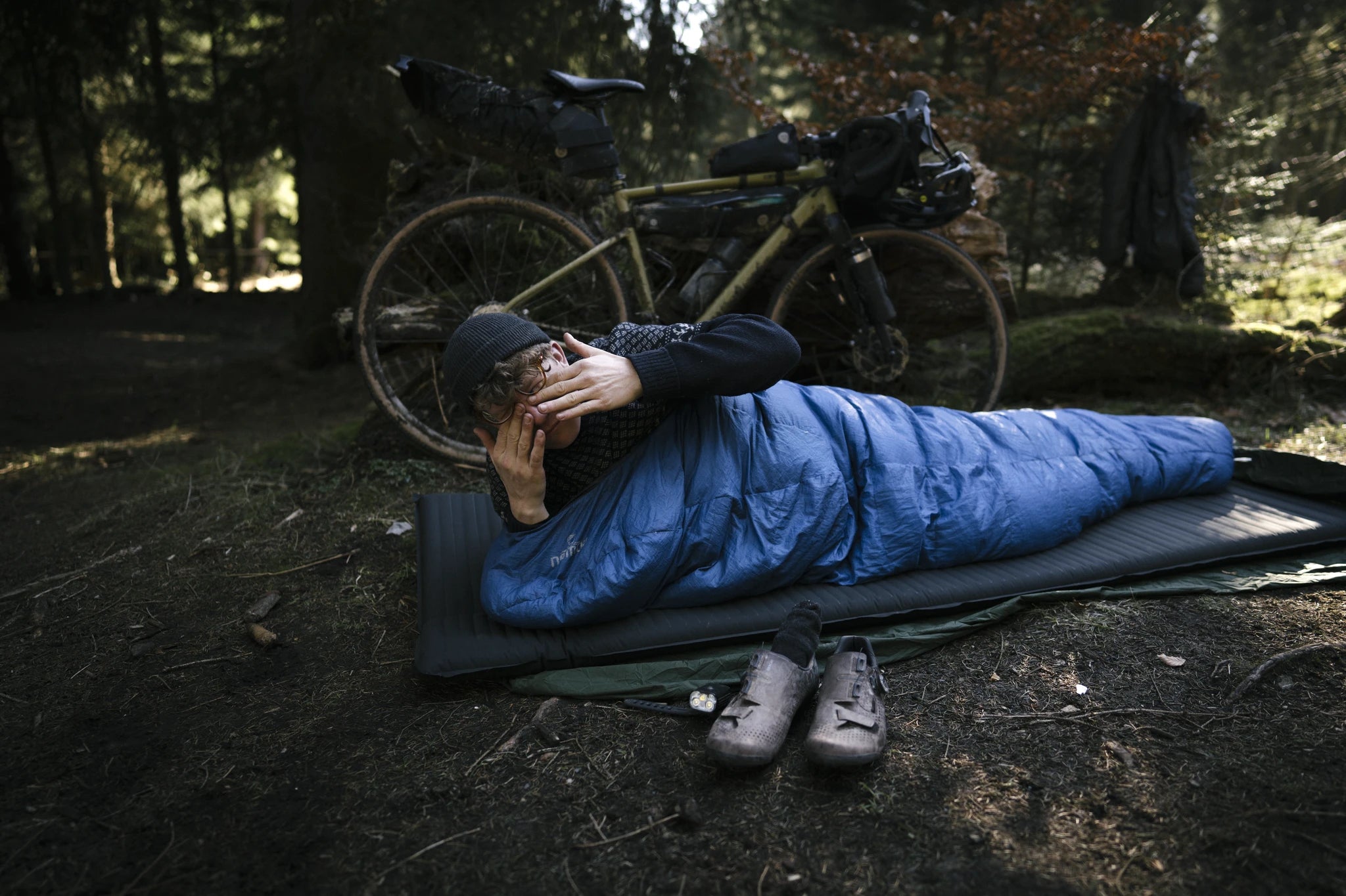Person im blauen Schlafsack auf Isomatte im Wald, neben einem beladenen Gravelbike und Fahrradschuhen. Outdoor-Camping.