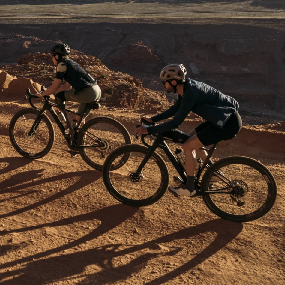 Zwei Personen fahren Gravelbikes auf einem sandigen Pfad in einer bergigen Landschaft, beide tragen Helme.