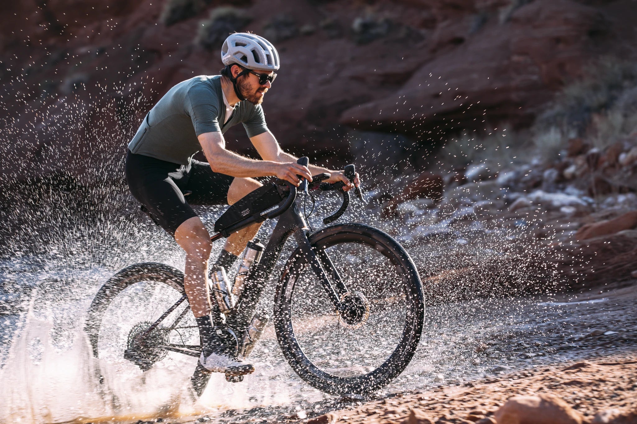 Mann auf Cannondale Gravelbike fährt durch Wasser in Wüstenlandschaft, trägt Helm und Radkleidung. Ideal für Outdoor-Sportler.
