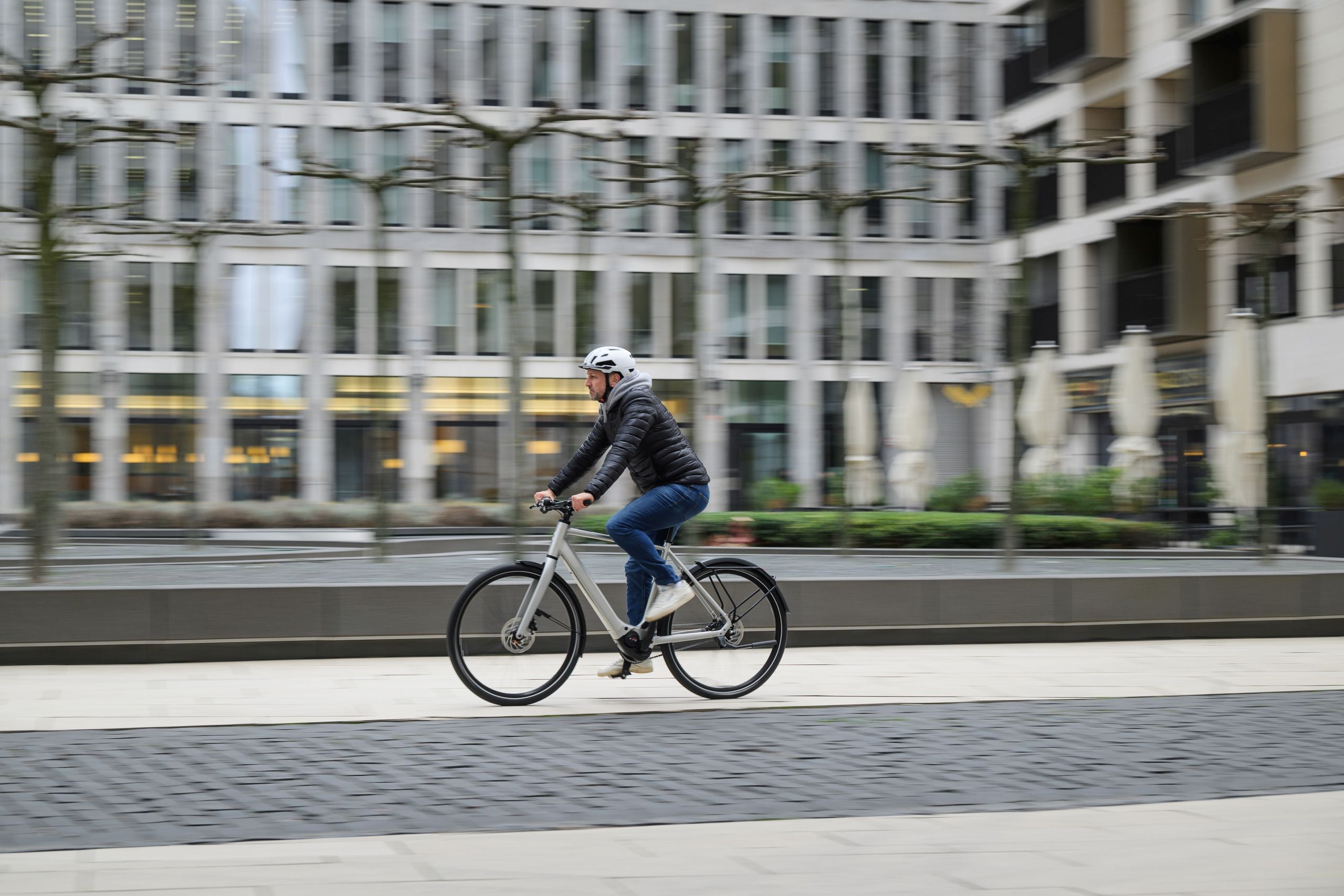 Person fährt Rad im urbanen Umfeld. Moderne Architektur im Hintergrund. Fahrrad und Sicherheitsausrüstung erkennbar.