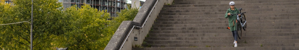 Person trägt Fahrrad eine lange Steintreppe hinunter, im Hintergrund grüne Bäume und ein Rohbau.