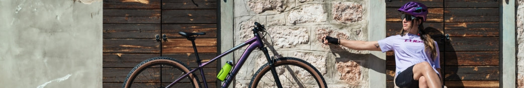 Frau mit lila Helm sitzt neben einem lila Gravelbike vor Holztür; grüner Flaschenhalter am Fahrradrahmen.