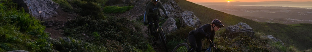 Zwei Mountainbiker fahren bei Sonnenuntergang auf einem felsigen Pfad in einer hügeligen Landschaft.
