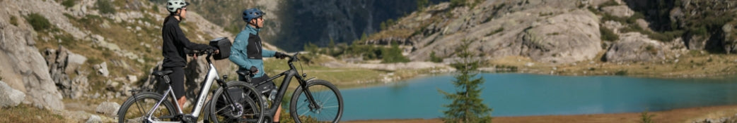 Zwei Personen mit E-Bikes stehen vor einem Bergsee in einer Berglandschaft, ausgerüstet mit Helmen und Outdoor-Kleidung.