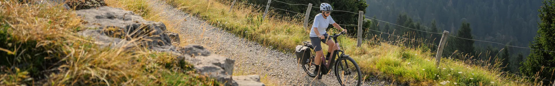 Person fährt auf einem Schotterweg in den Bergen ein E-Bike mit Gepäcktaschen, Fahrradhelm und sportlicher Bekleidung.