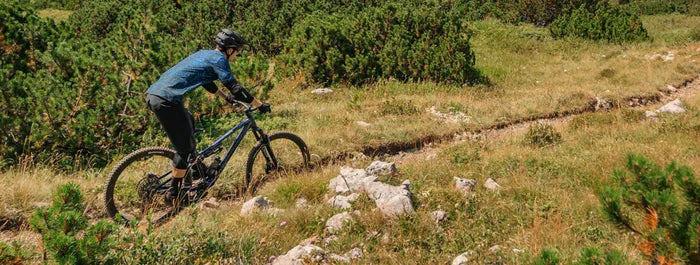 Mountainbikerin auf unbefestigtem Pfad in bergiger Landschaft, mit Schutzkleidung und Helm, ideal für Outdoor-Aktivitäten.