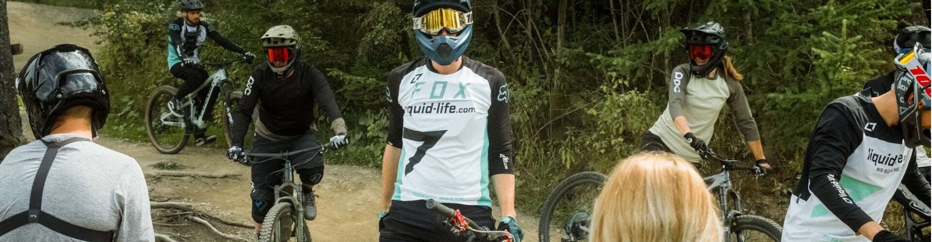 Mountainbiker in Schutzkleidung und Helmen, teils mit FOX- und POC-Logos, fahren auf Waldstrecke.