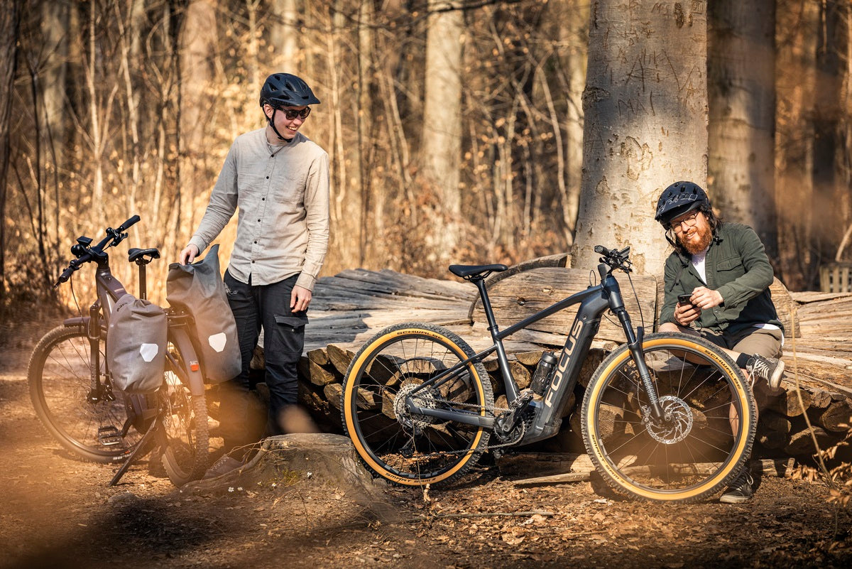 Zwei Männer mit Focus E-Bikes im Wald, einer mit Handy, der andere mit Radpacktaschen, tragen Helme.