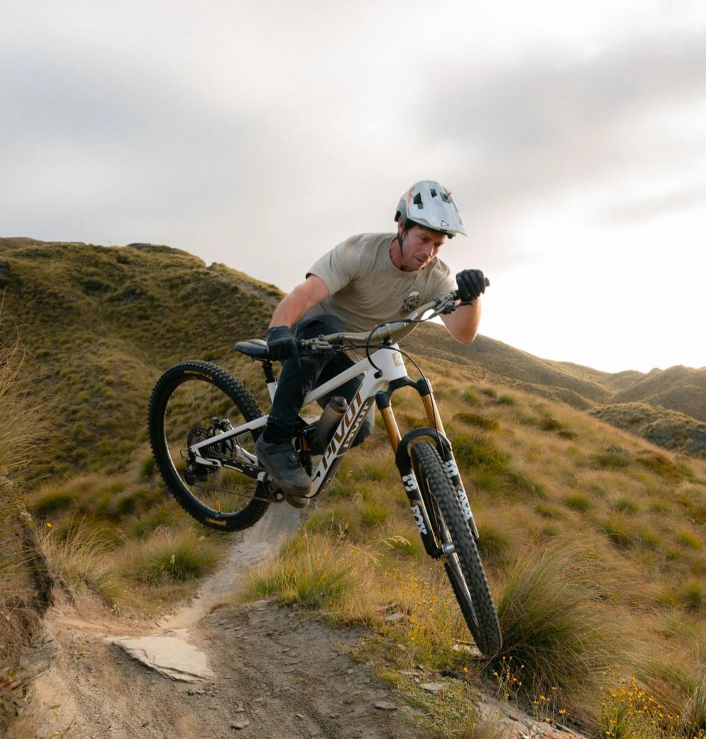 Mountainbiker auf einem Pivot Bike meistert einen Sprung auf einem schmalen Pfad in einer hügeligen Landschaft.