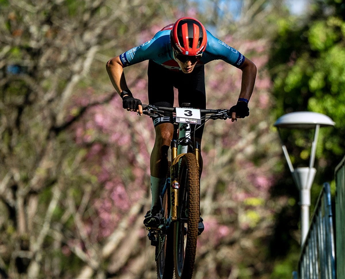 Mountainbiker auf Cube-Rad in Aktion, getragenes Outfit für Wettkämpfe, im Hintergrund Bäume und Blumen.