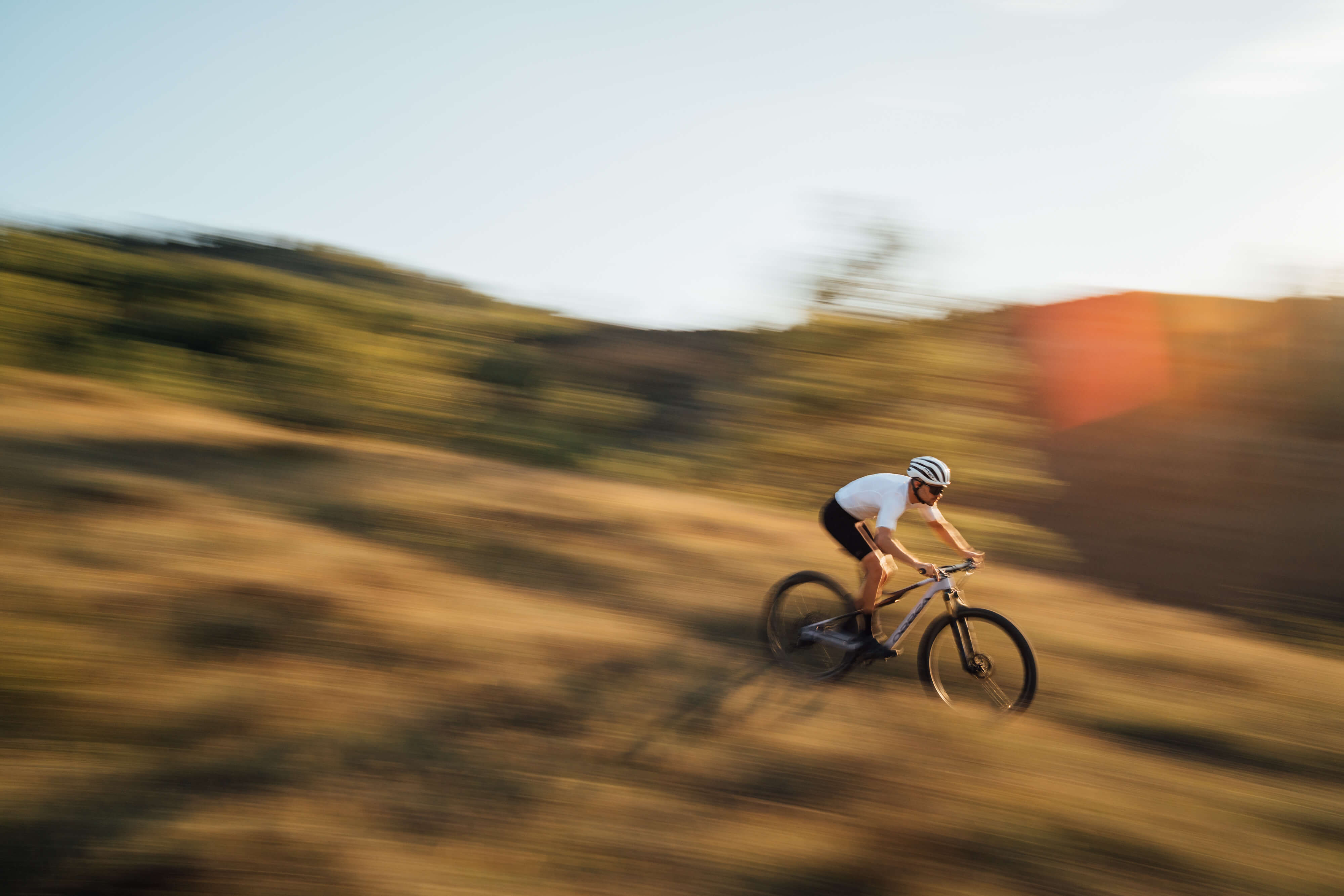 Radfahrer auf einem Orbea Mountainbike fährt schnell durch hügelige Landschaft bei Sonnenuntergang.