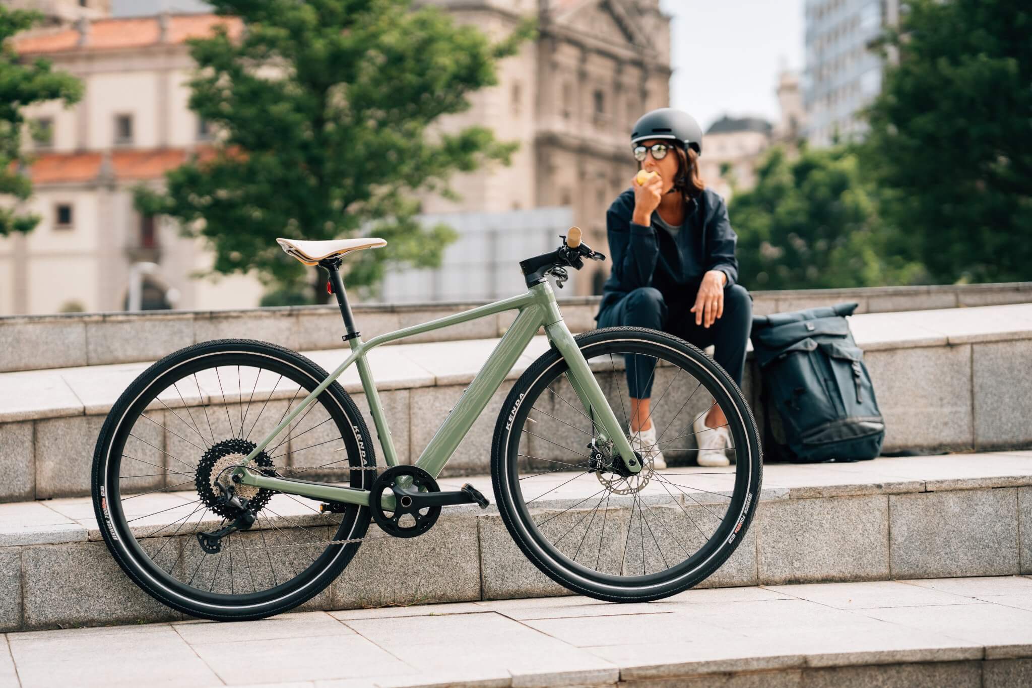 Grünes Orbea Vibe Rad auf Stufen, Frau mit Helm isst und sitzt daneben, Stadthintergrund. Ideal für urbane Pendler.
