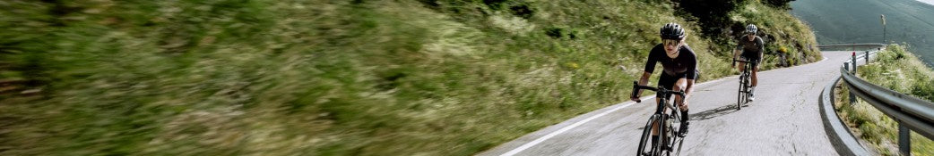 Zwei Radfahrer mit Helmen fahren auf einer kurvigen Bergstraße entlang, umgeben von grüner Landschaft.