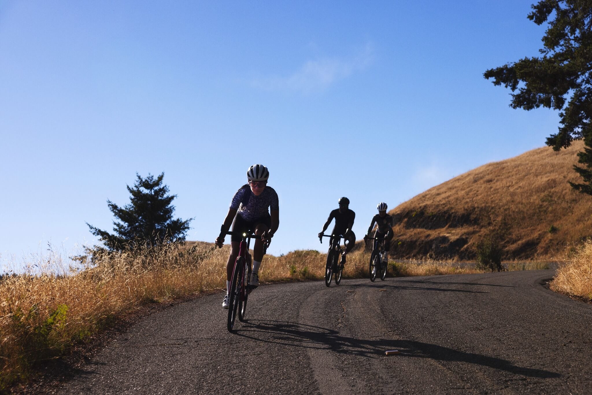Drei Radfahrer auf einer kurvigen Landstraße, bei sonnigem Wetter und hügeliger Landschaft.
