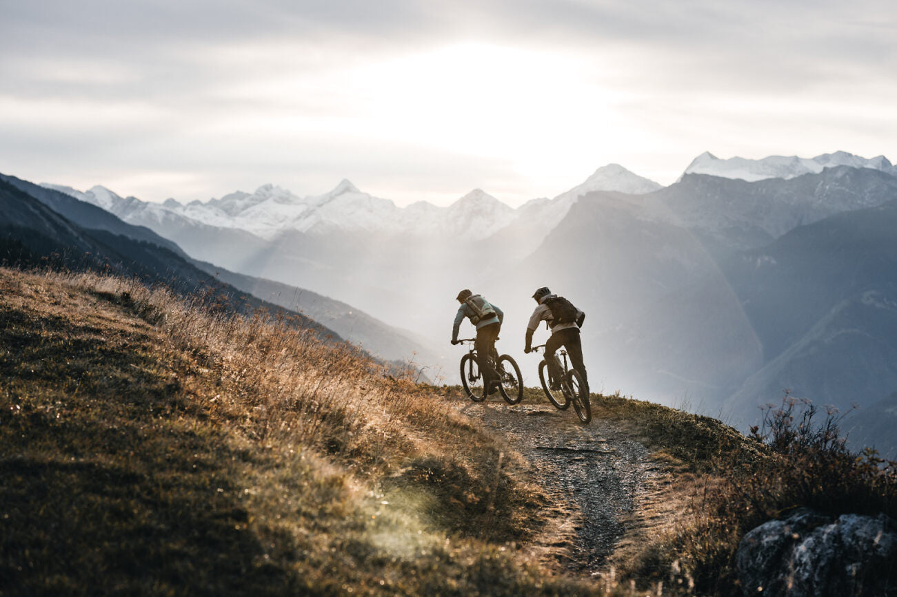 Zwei Mountainbiker fahren abends auf einem Gebirgspfad mit Specialized E-Mountainbikes in alpiner Landschaft.