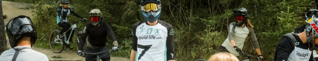 Mountainbiker in Schutzkleidung und Helmen in einem Wald, tragen Trikots von "liquid-life.com", bereit für Abfahrt.