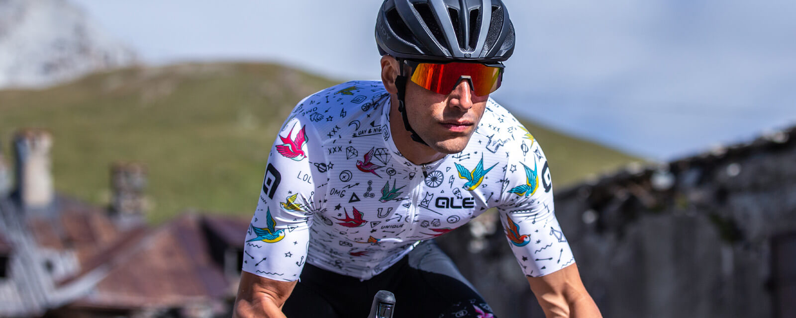 Radsportler in Ale-Radtrikot mit buntem Muster, trägt Helm und Sonnenbrille, im Freien auf dem Fahrrad.