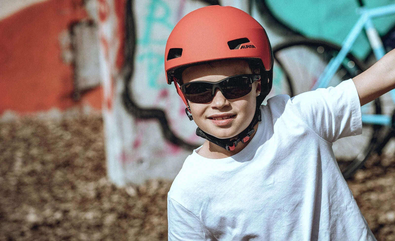 Kind mit rotem Alpina-Fahrradhelm und Sonnenbrille, lächelnd vor bunter Wand und Fahrrad im Hintergrund.