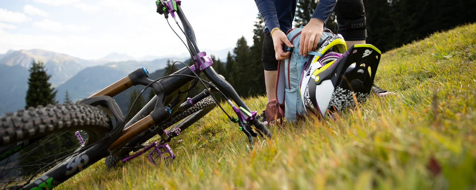Fahrrad und Person mit Rucksack auf Wiese. Ein Mountainbike-Helm wird aus dem Rucksack geholt.