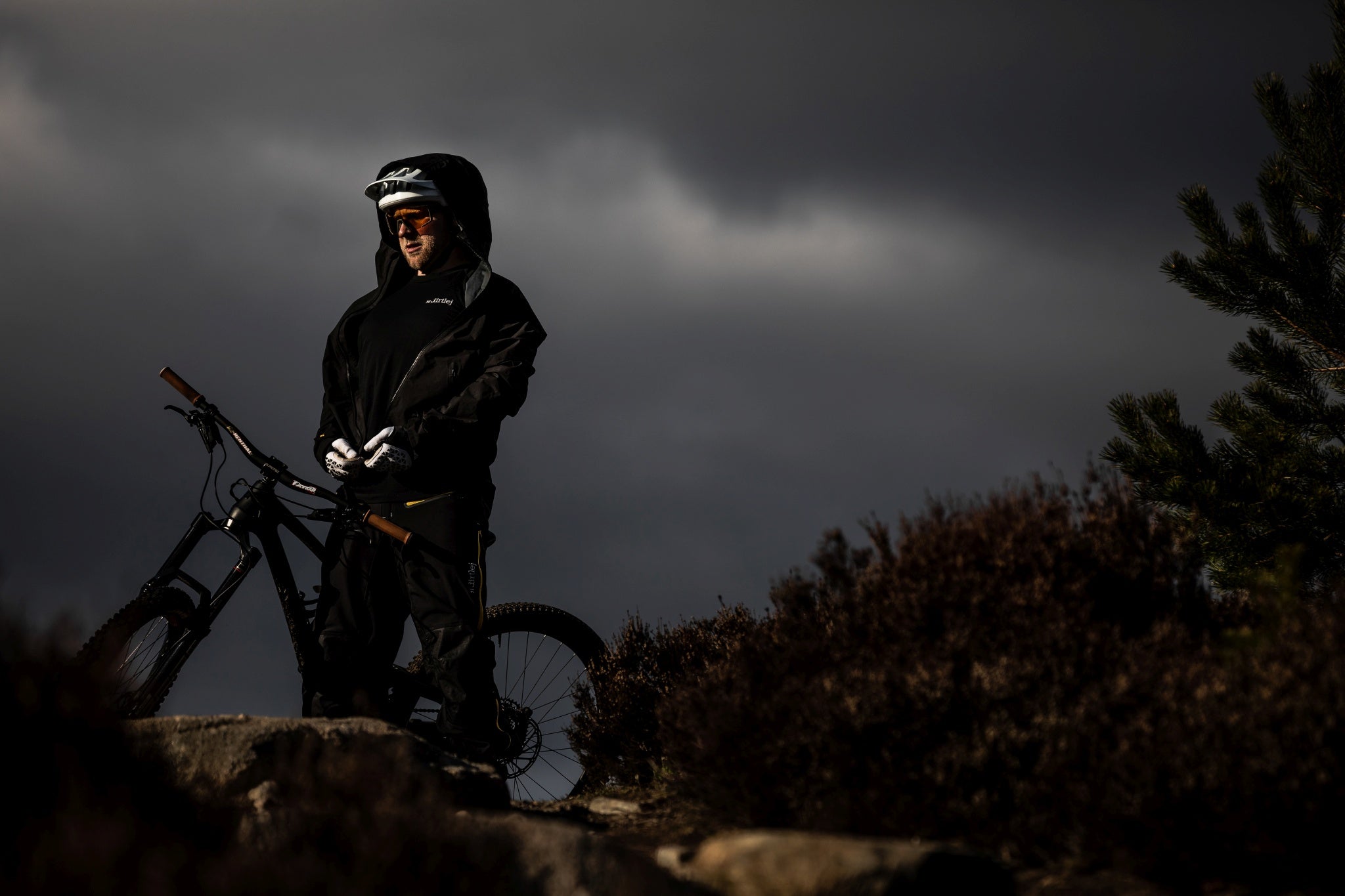 Person mit schwarzer Mountainbike-Ausrüstung und Helm neben einem Mountainbike in rauem Gelände bei bewölktem Himmel.