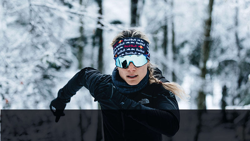 Läuferin im verschneiten Wald mit Red Bull Stirnband, Sonnenbrille und The North Face Jacke beim Winterlauf.