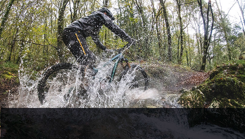 Mountainbiker fährt durch Wasser in einem Wald; Kleidung aus der Kollektion "Regen-Wetterschutz".