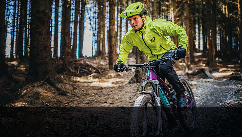 Mann auf Mountainbike im Wald, trägt grüne Regenjacke und Helm, Kollektion "Regen Wetterschutz".