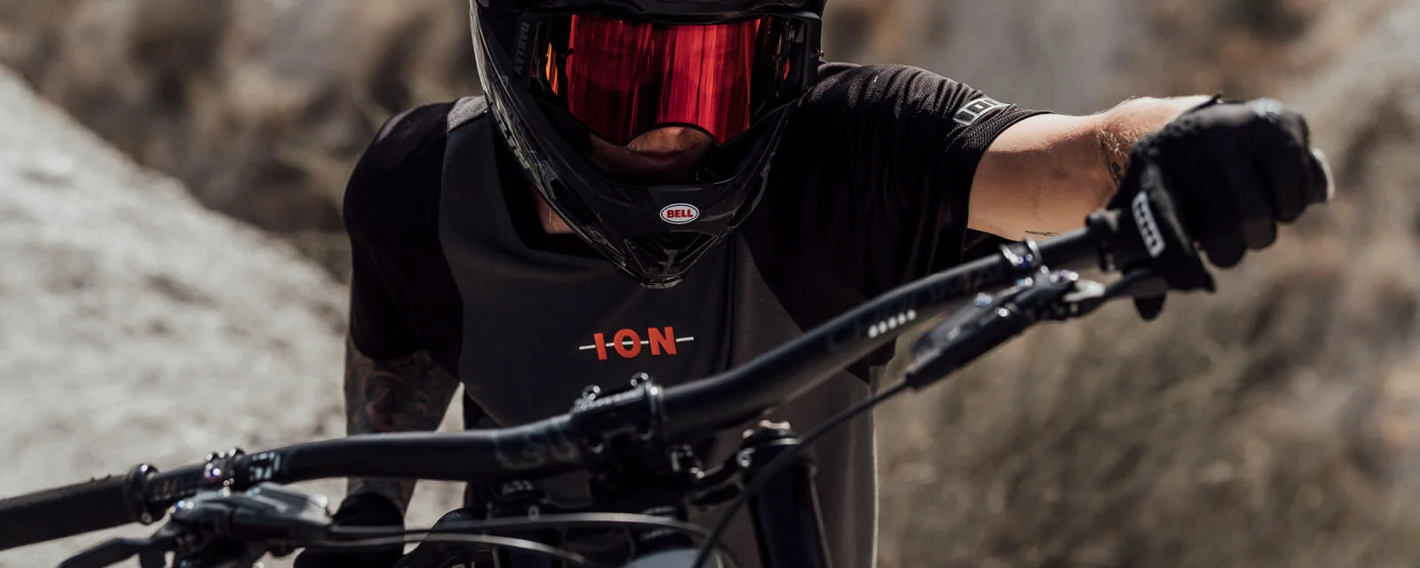 Mountainbiker mit Helm und Brille, trägt ein schwarzes ION-Shirt, auf einem Mountainbike in felsiger Landschaft.