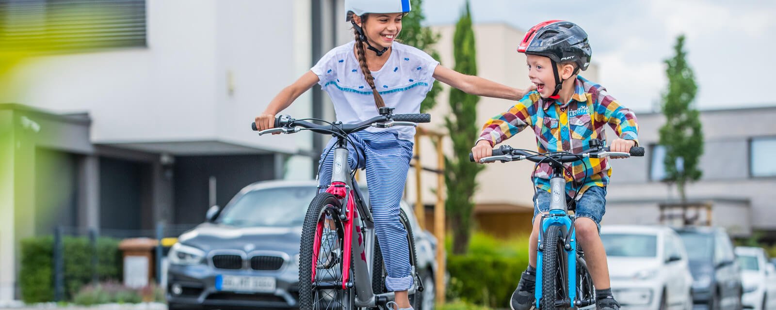 Zwei Kinder auf Fahrrädern mit Helmen lächeln draußen in einer Wohnsiedlung; Junge auf PUKY-Fahrrad.