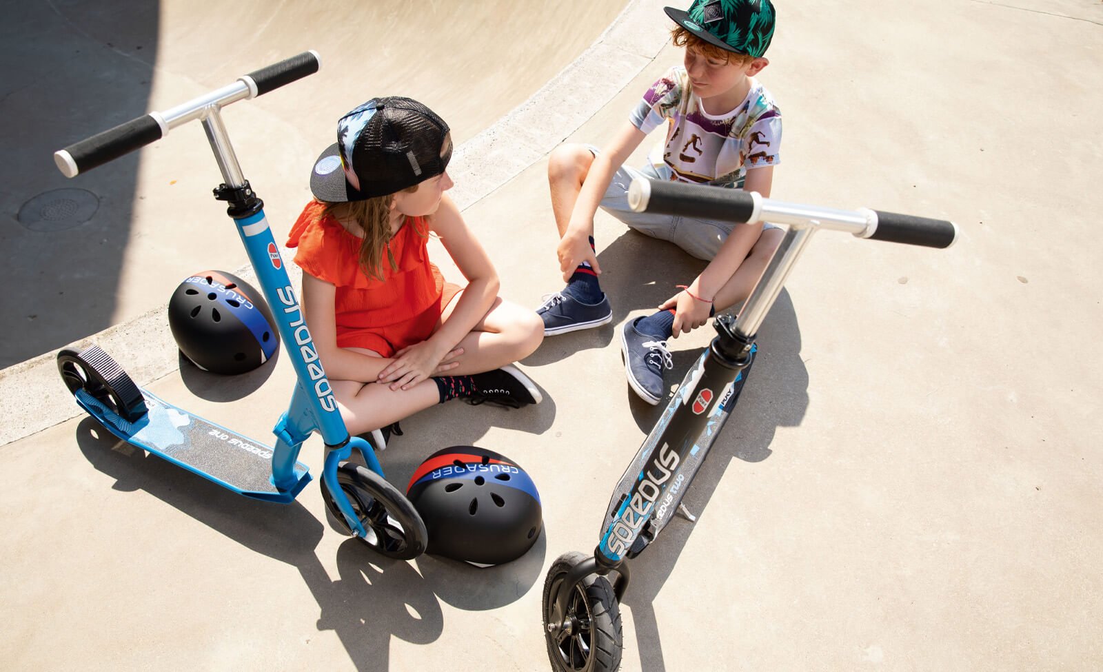 Kinder sitzen neben PUKY Scooters und Helmen auf Betonboden; Einsatz im Freizeitbereich für Kinder.