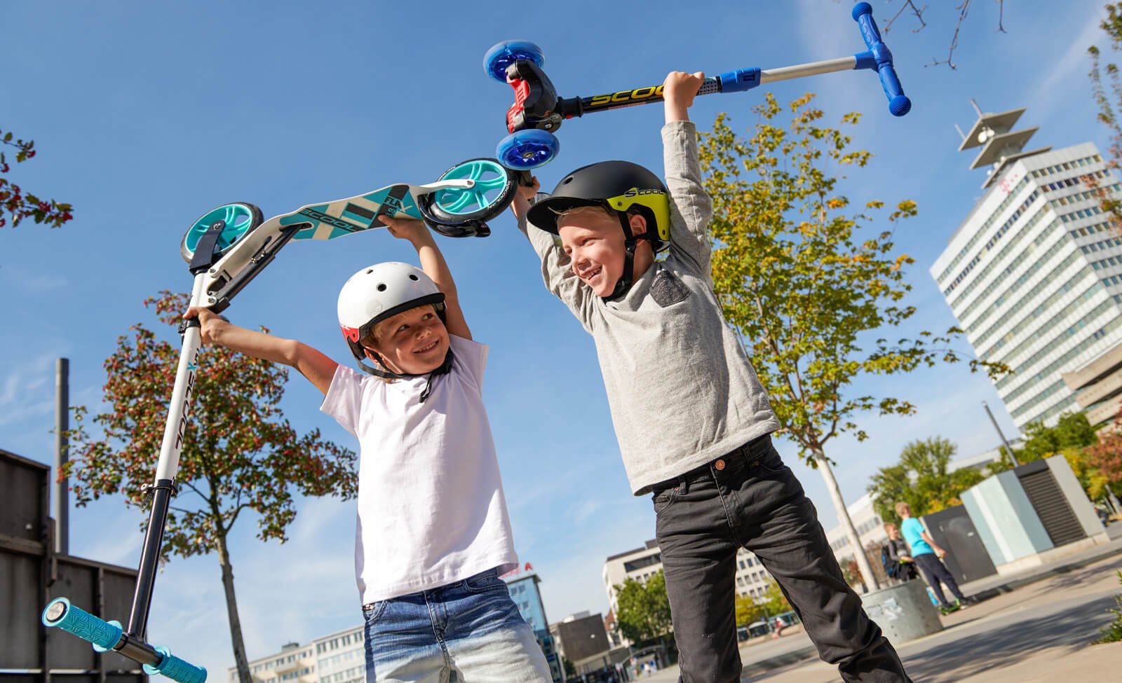 Zwei Kinder in Helmen halten begeistert zwei Scooter von Scool in die Luft, draußen bei sonnigem Wetter.